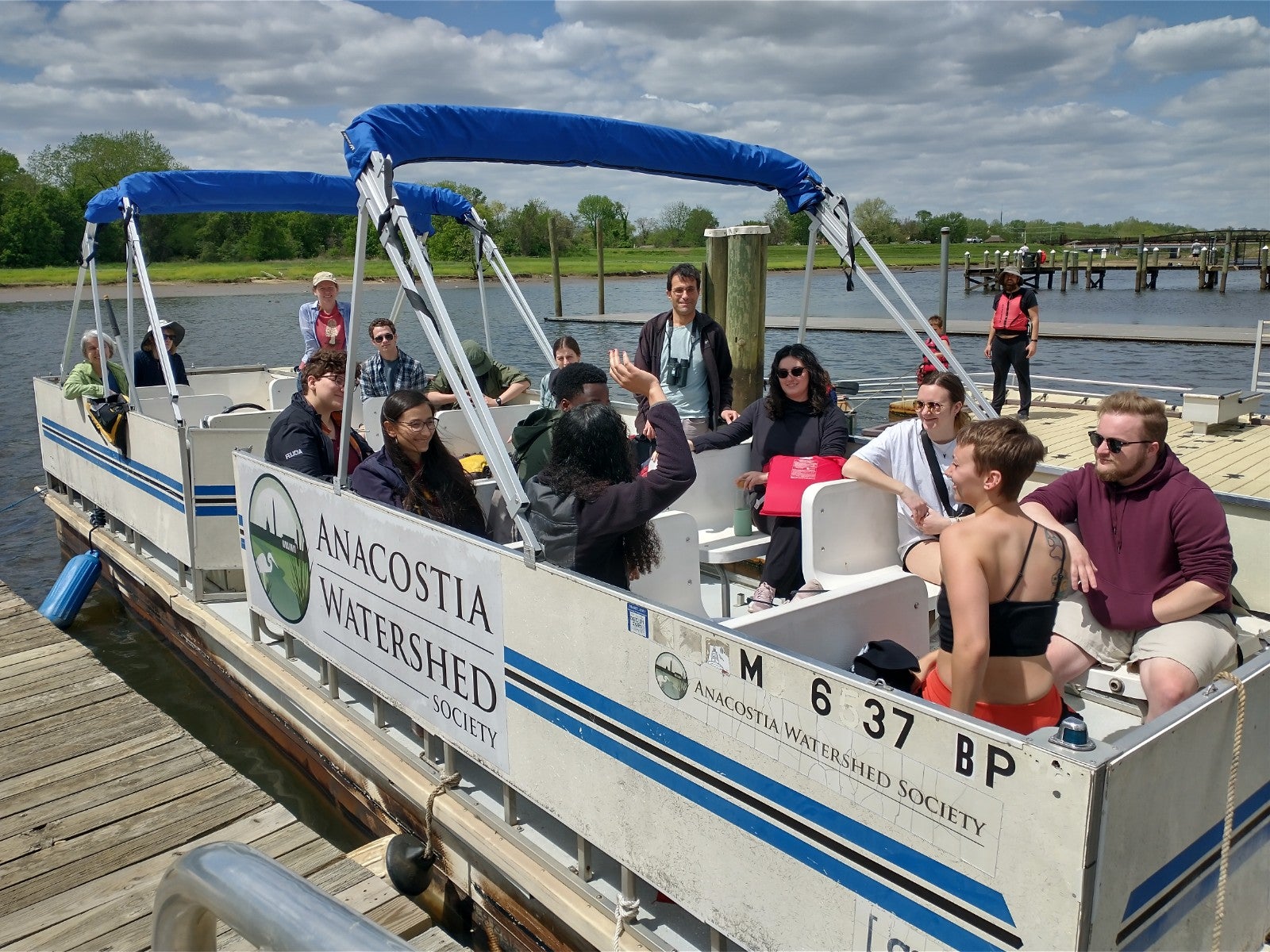 Students in a docked boat on the river waiting to go on the tour. It is a cloudy but bright day and the boat has the Anacostia Watershed Society logo on it