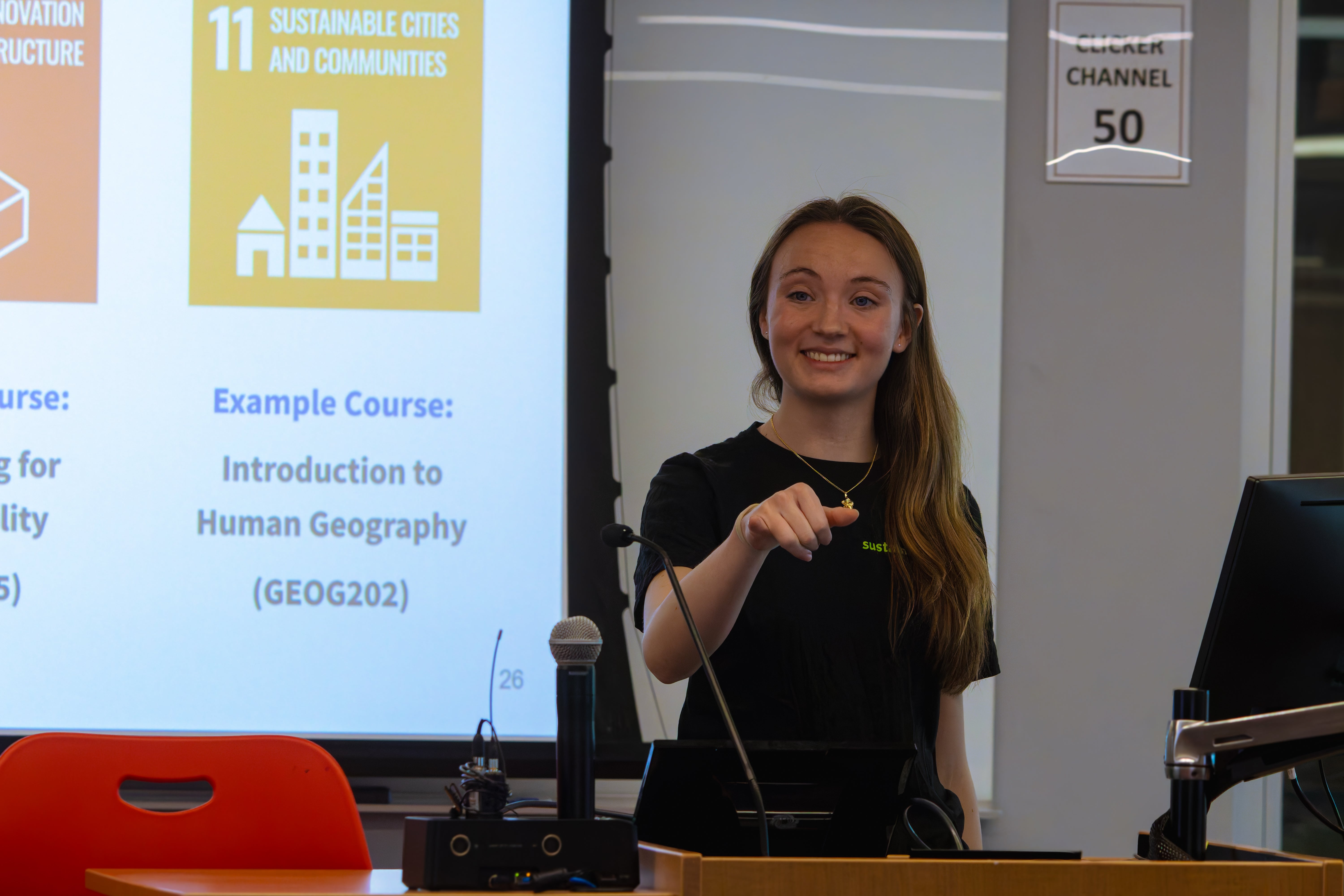 Student wearing SustainableUMD t-shirt smiles while leading a presentation. Screen behind shows one of the UN Sustainable Development Goals and an example course (Intro to Human Geography)