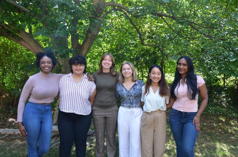 A group of six students dressed in business casual pose for a photo in front of a tree
