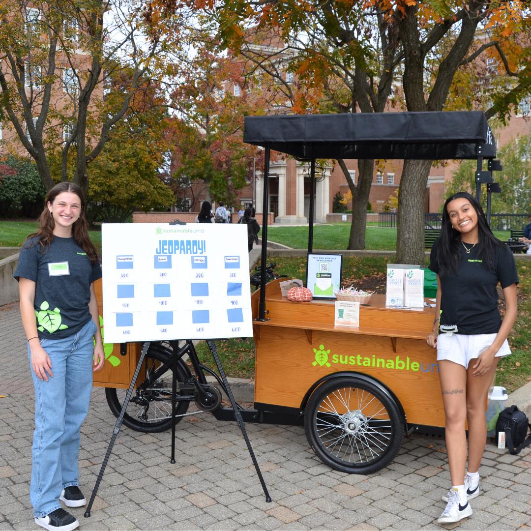 Two SustainableUMD Outreach Ambassadors pose in front of a jeopardy board at the SustainableUMD Outreach Bike.