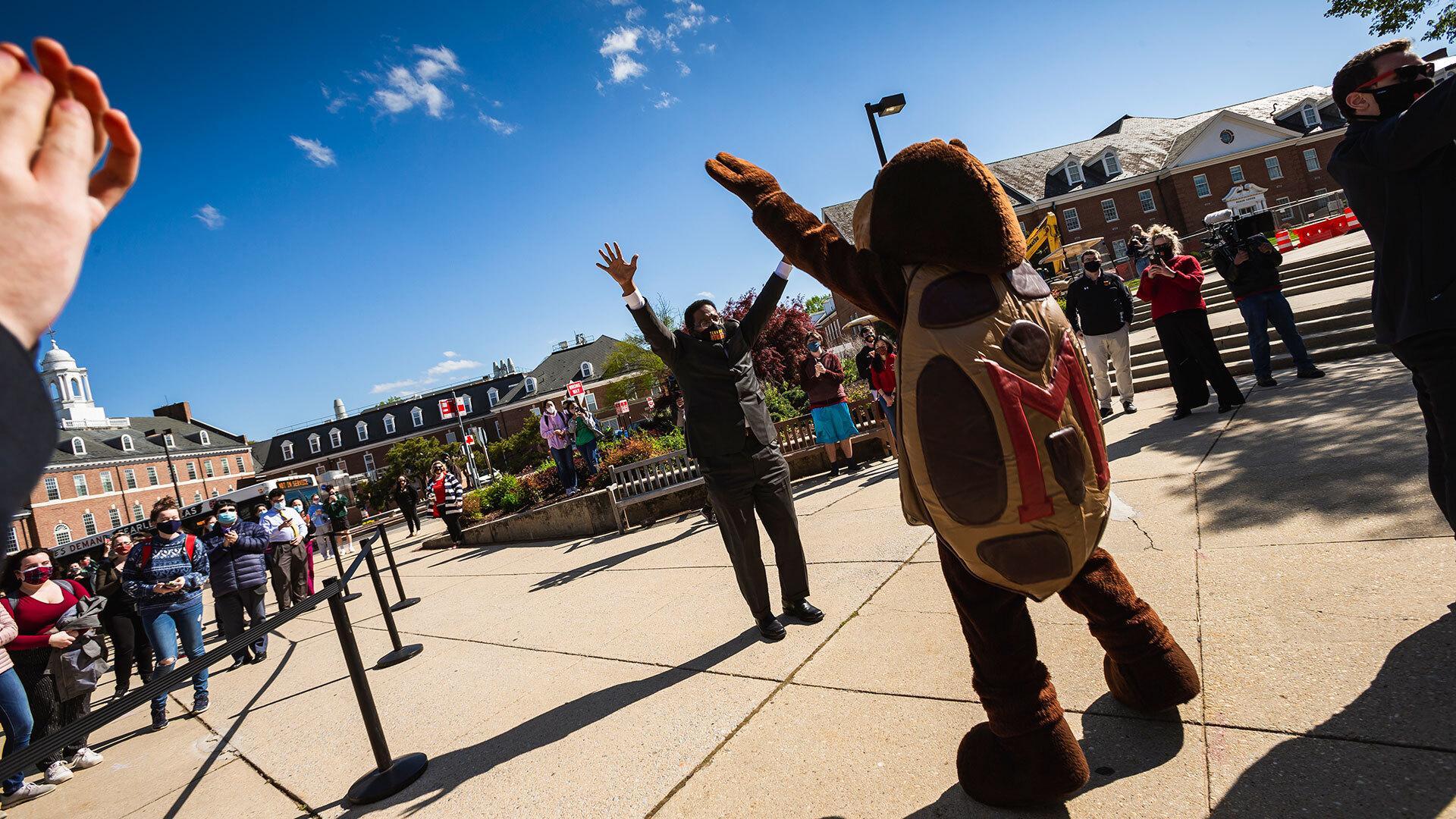 Pines and Testudo with arms up on campus