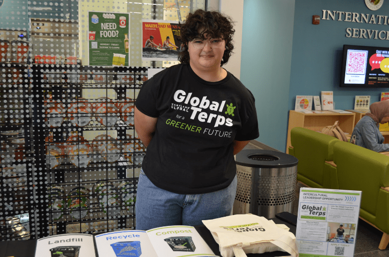 A student wearing a black Global Terps for a Greener Future shirt with a green leaf turtle poses in front of a table with a waste sorting activity