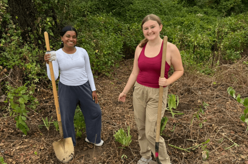 Two students stand in a patch of dirt and plants holding shovels