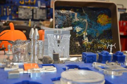 A spread of various 3D printed materials (including mini, blue testudo statues) and metal objects made from molds (including a solid M and a Yoda figure)